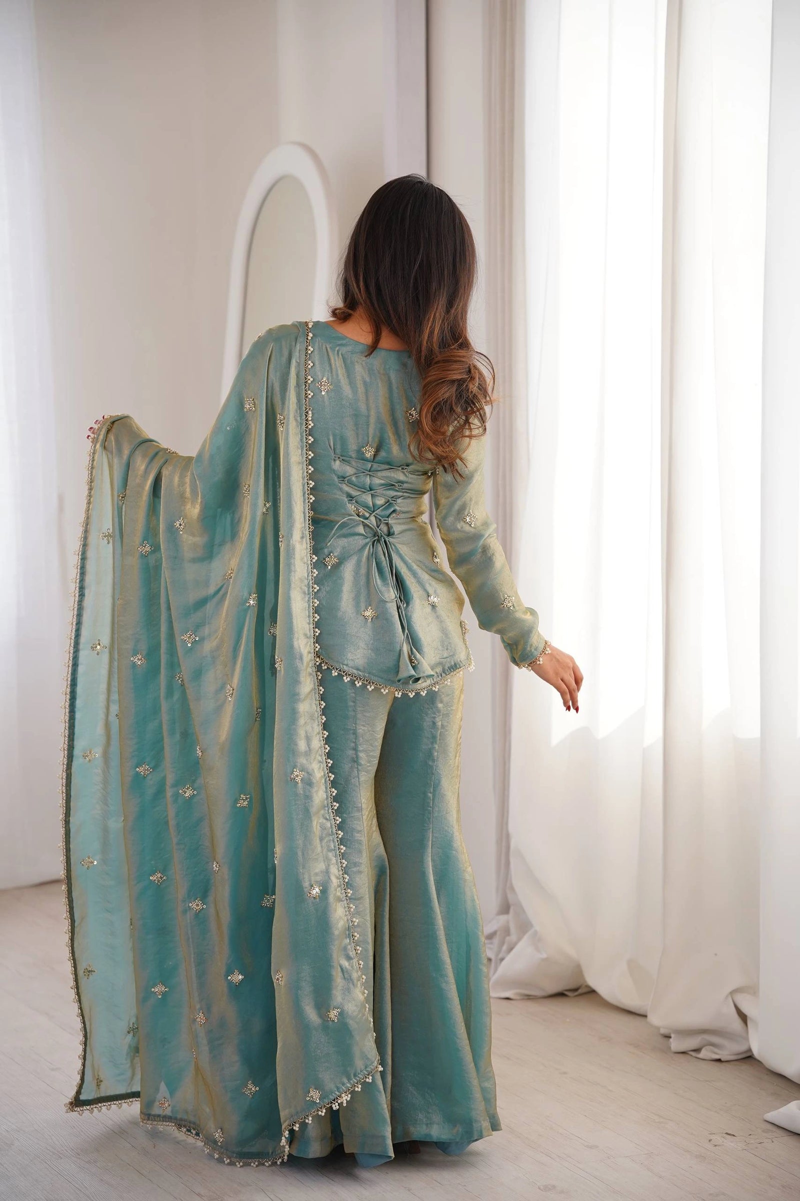 Woman wearing a teal traditional outfit with intricate designs, standing in a room with white curtains.
