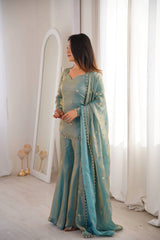 Woman in a light blue traditional outfit standing in a room with white curtains and a mirror.