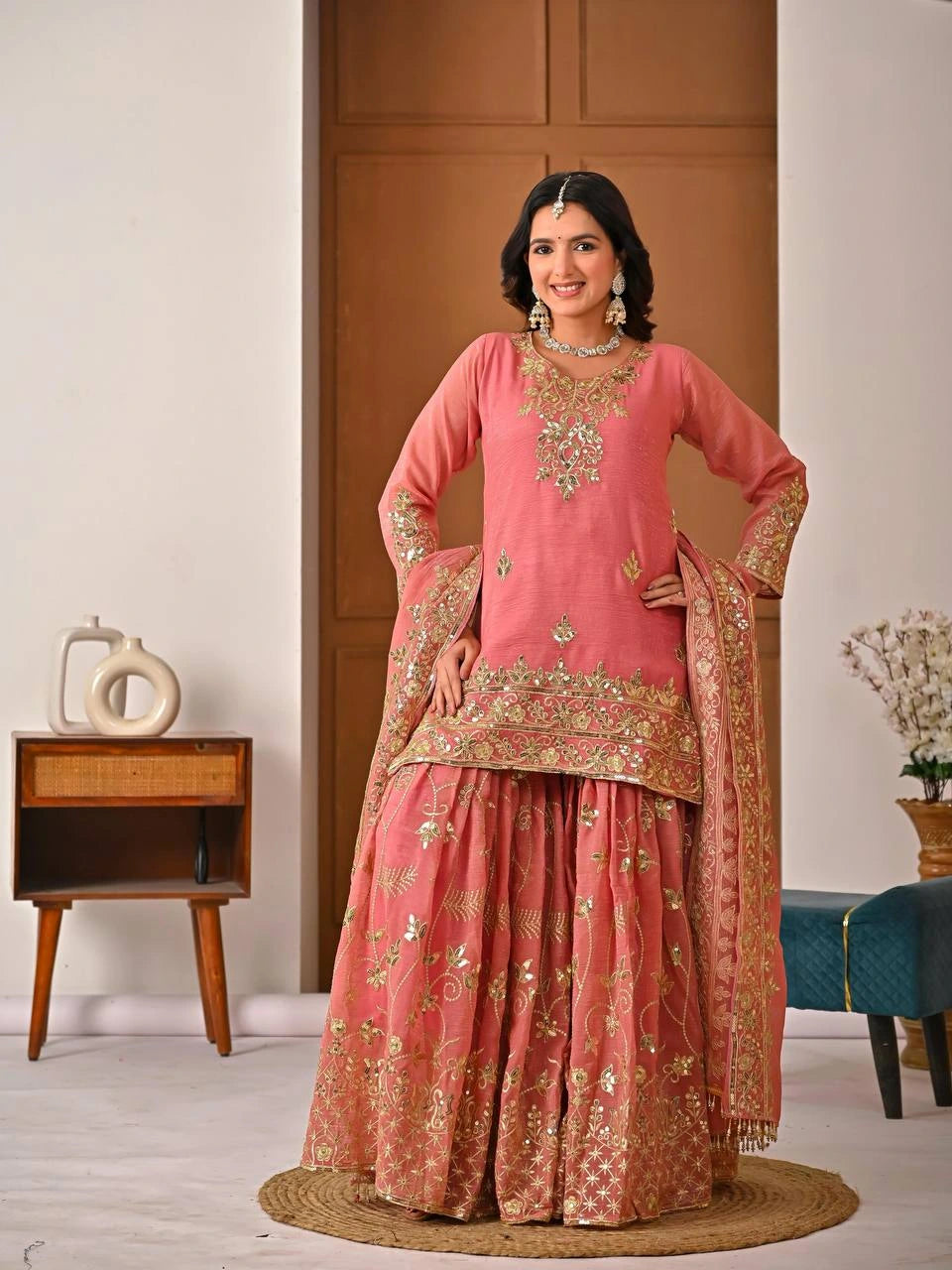 Woman in a traditional pink and gold embroidered outfit standing in a room with wooden furniture.
