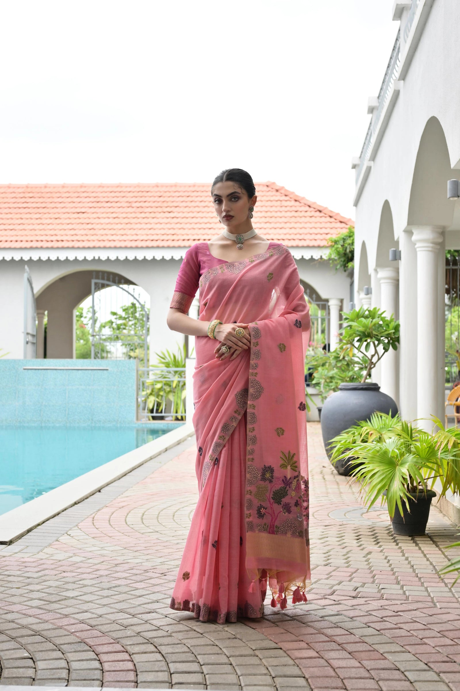 Woman in a pink saree standing by a poolside with a white building and greenery in the background.