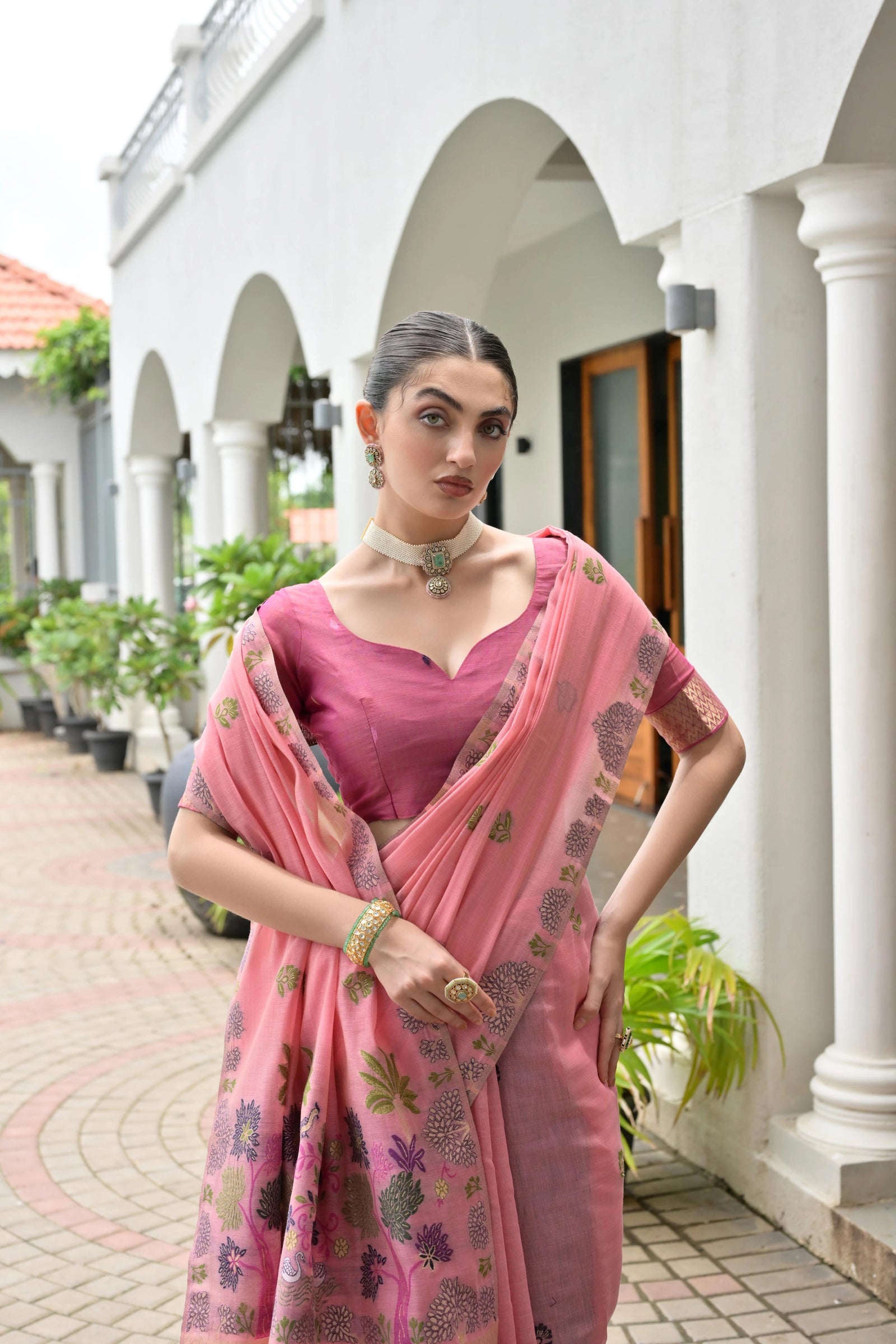 Woman in a pink saree standing in front of a white building with arches.