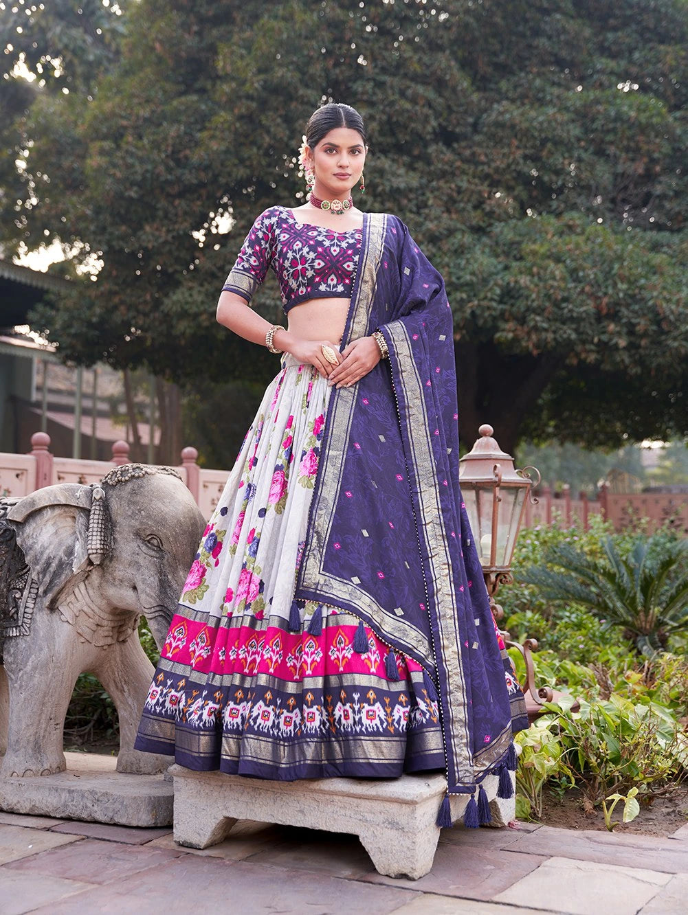 Woman in a traditional purple and pink lehenga standing outdoors with greenery in the background