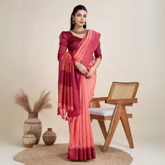 A woman models a striking two-toned Raw Silk Saree. The main body is Coral Pink, and the pallu/border contrast dramatically in Deep Maroon/Wine Red. The saree features traditional temple weaving and horizontal stripe patterns on the pallu. She wears a long-sleeve maroon blouse and accessorizes with a chunky dark gemstone necklace, posing indoors.