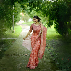 A woman models a Terracotta Red Soft Cotton Saree featuring an all-over digital block print of traditional motifs. She wears a printed red blouse and silver jhumka earrings, posing on an outdoor path framed by greenery.