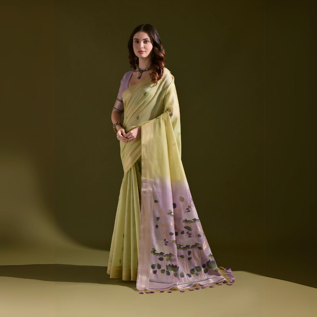 A woman models a Soft Muga Cotton Saree. The saree's body is soft light green with a gold Zari border. The pallu transitions into a light purple color featuring large, artistic woven motifs in green and purple. She wears a lavender blouse and a dark choker necklace.