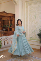 Woman in a light blue traditional outfit standing in an ornate room with wooden furniture and decorative wall.