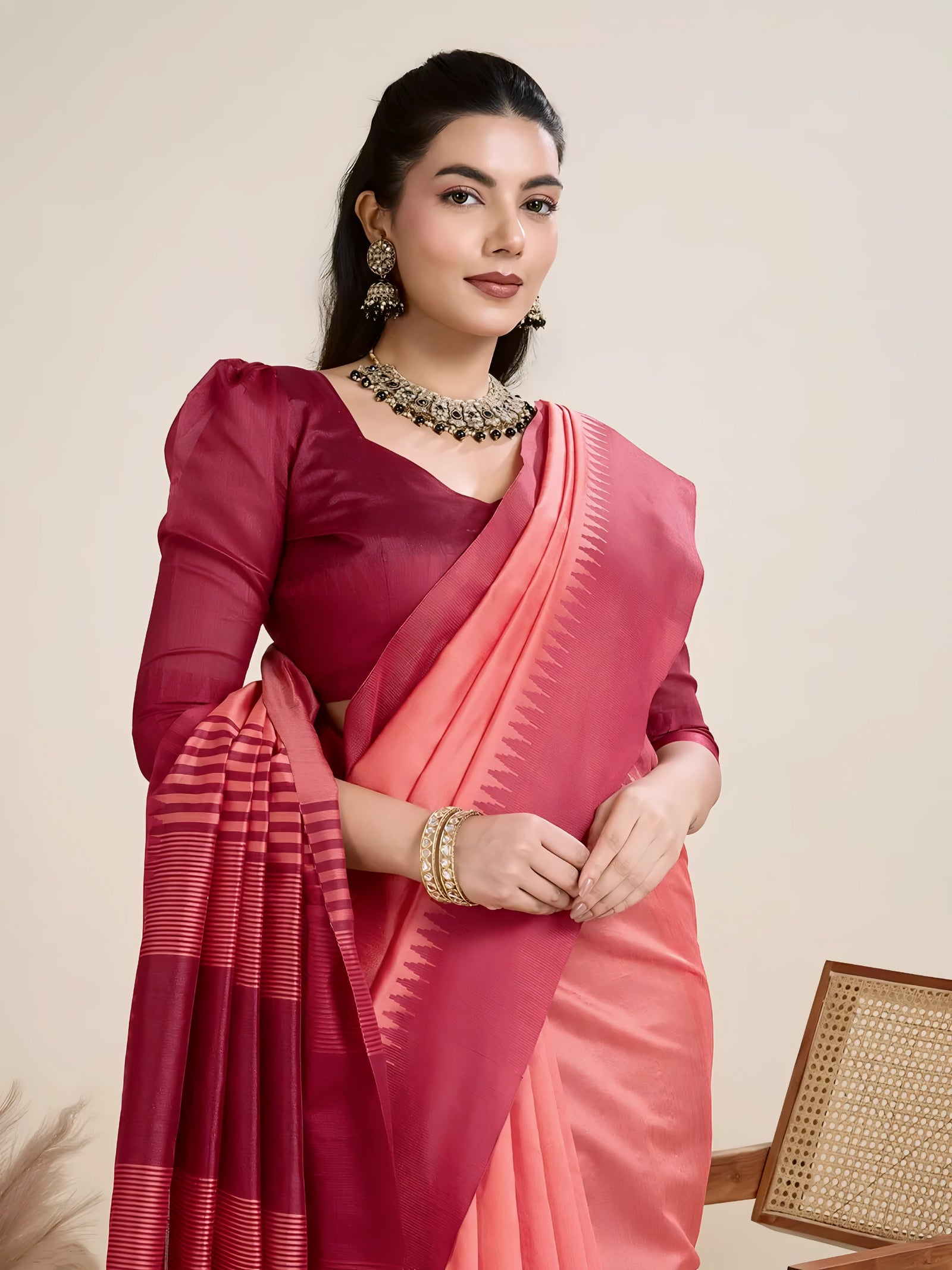 A woman models a striking two-toned Raw Silk Saree. The main body is Coral Pink, and the pallu/border contrast dramatically in Deep Maroon/Wine Red. The saree features traditional temple weaving and horizontal stripe patterns on the pallu. She wears a long-sleeve maroon blouse and accessorizes with a chunky dark gemstone necklace, posing indoors.