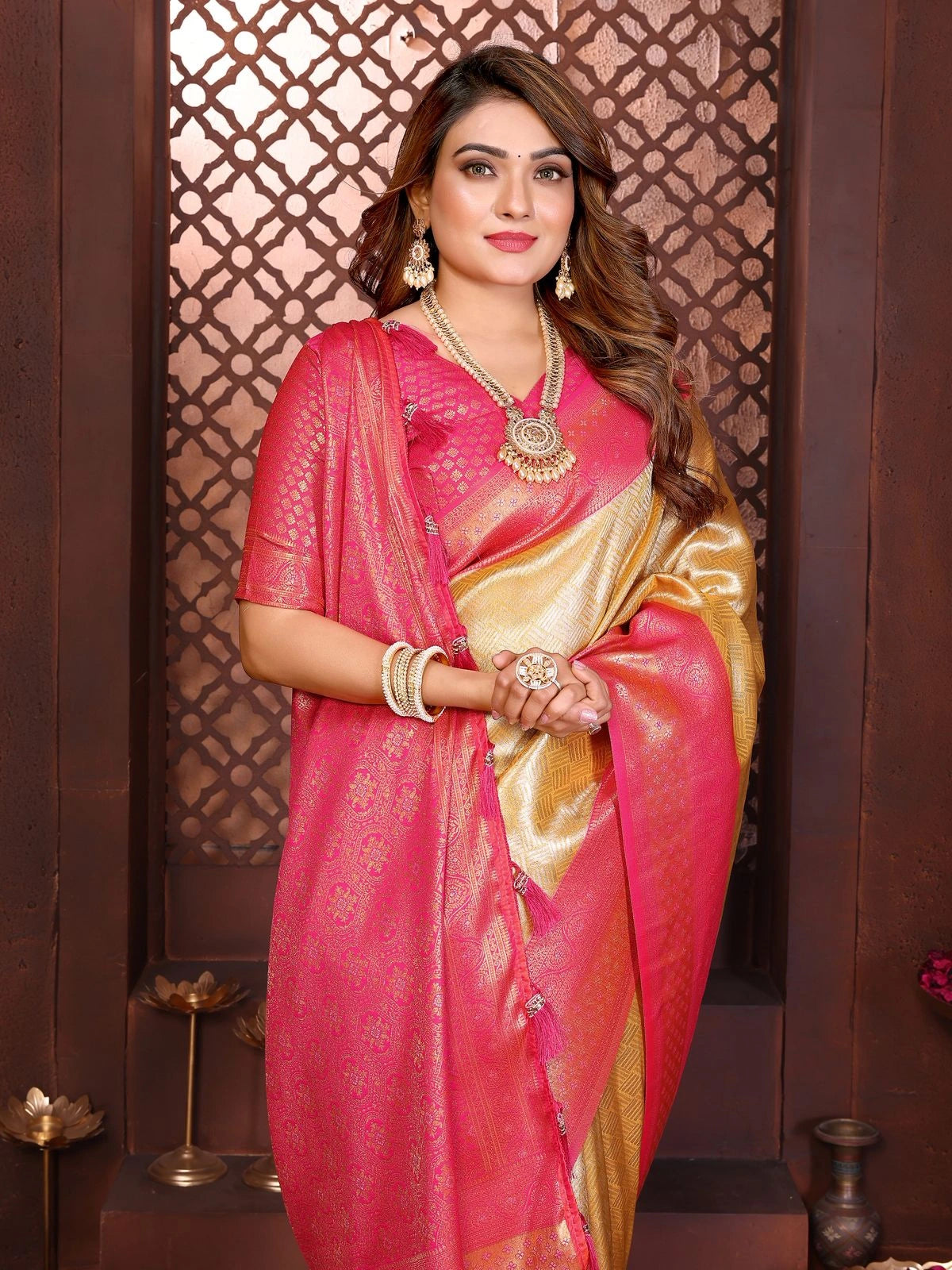 Woman wearing a pink and gold saree with jewelry against a decorative wall.