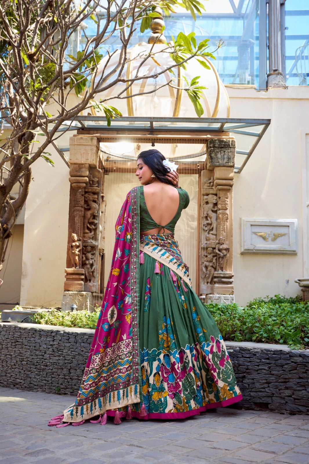 Woman in a traditional green and multicolored saree standing outdoors with decorative elements in the background.