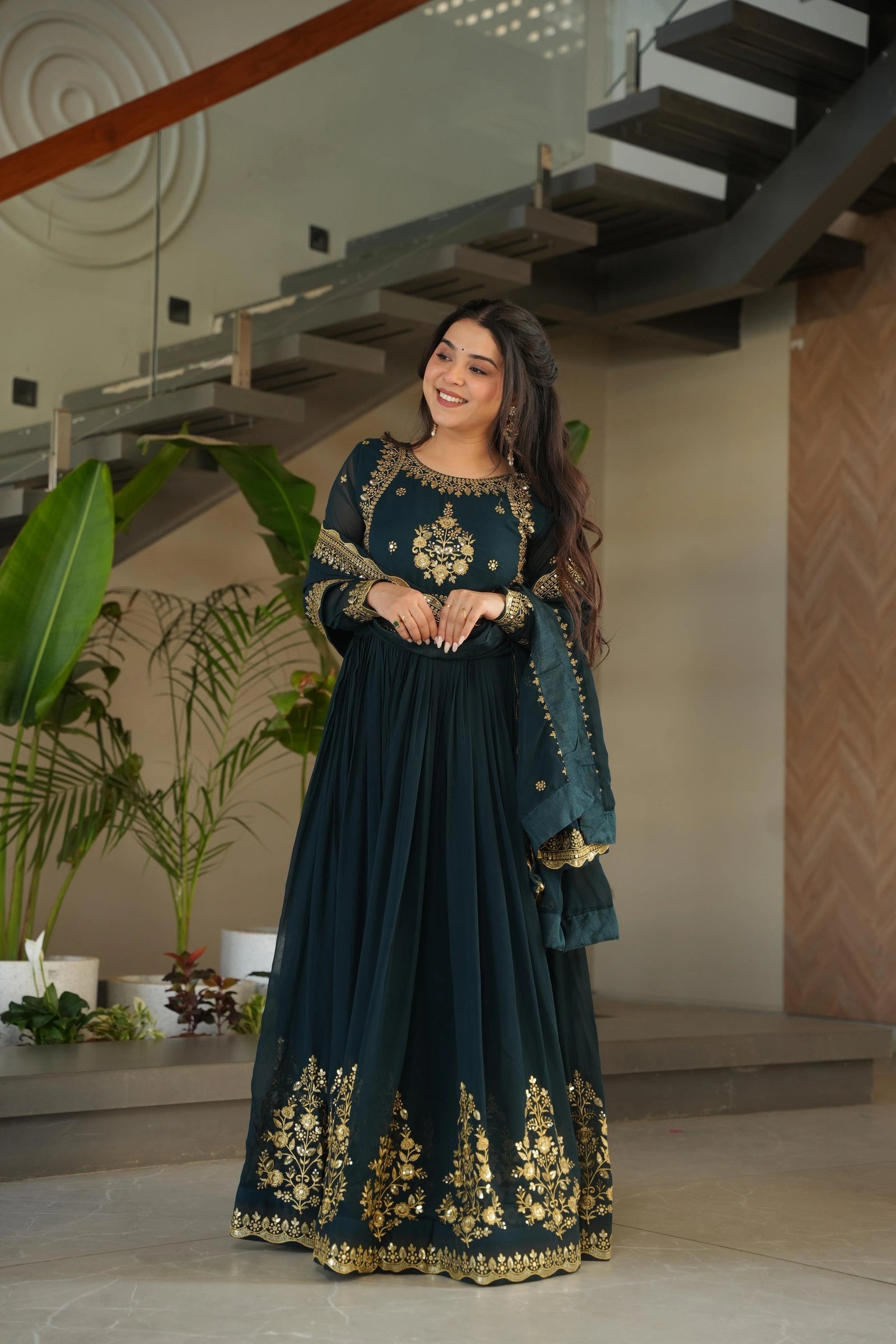 Woman in a dark green traditional outfit with gold embroidery standing indoors.