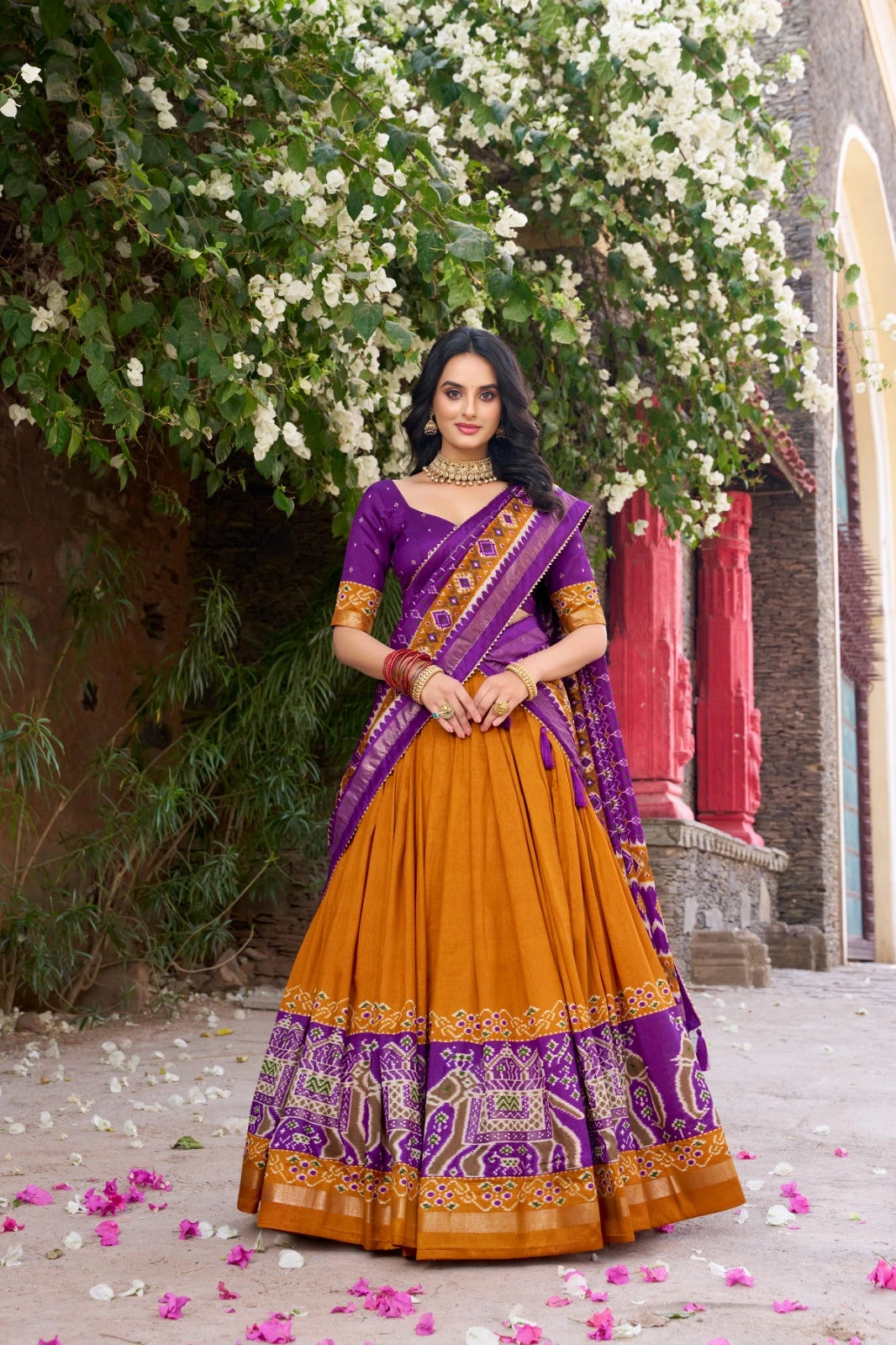 Woman in a traditional purple and orange saree standing under a tree with white flowers.