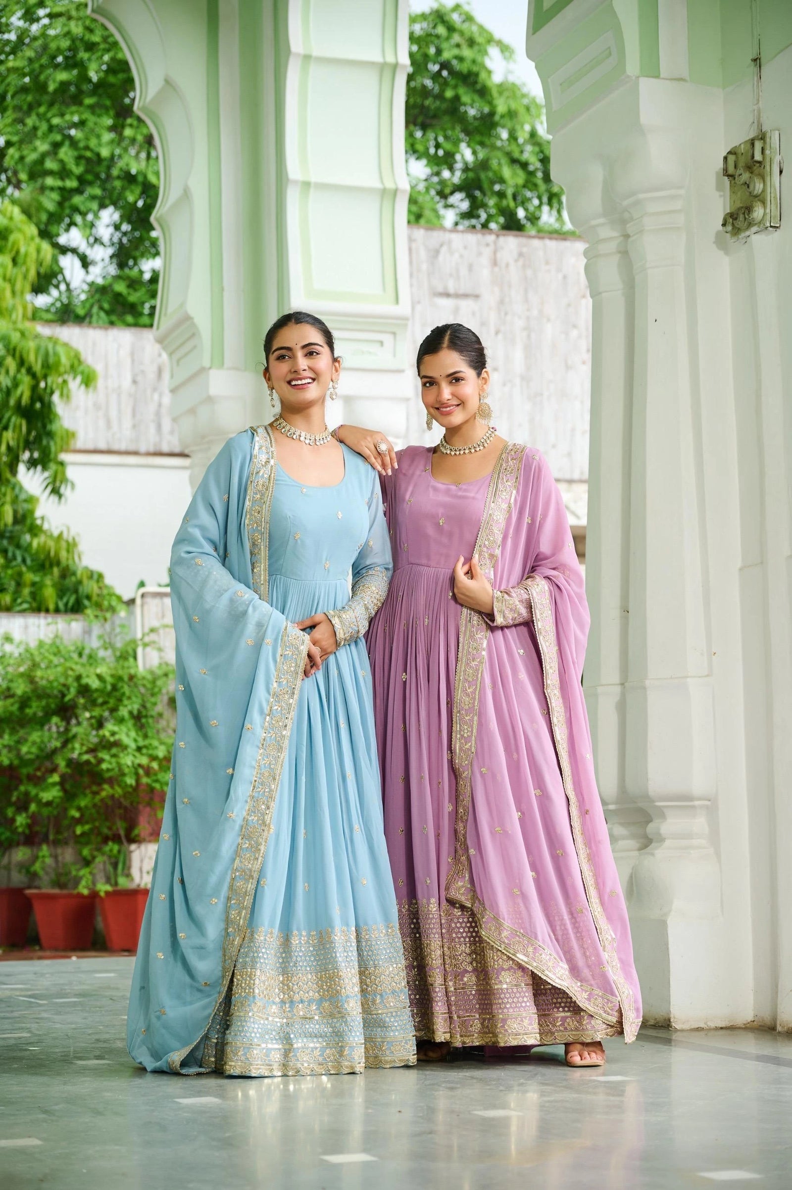 Two women in traditional light blue and pink sarees standing in front of a white architectural structure.