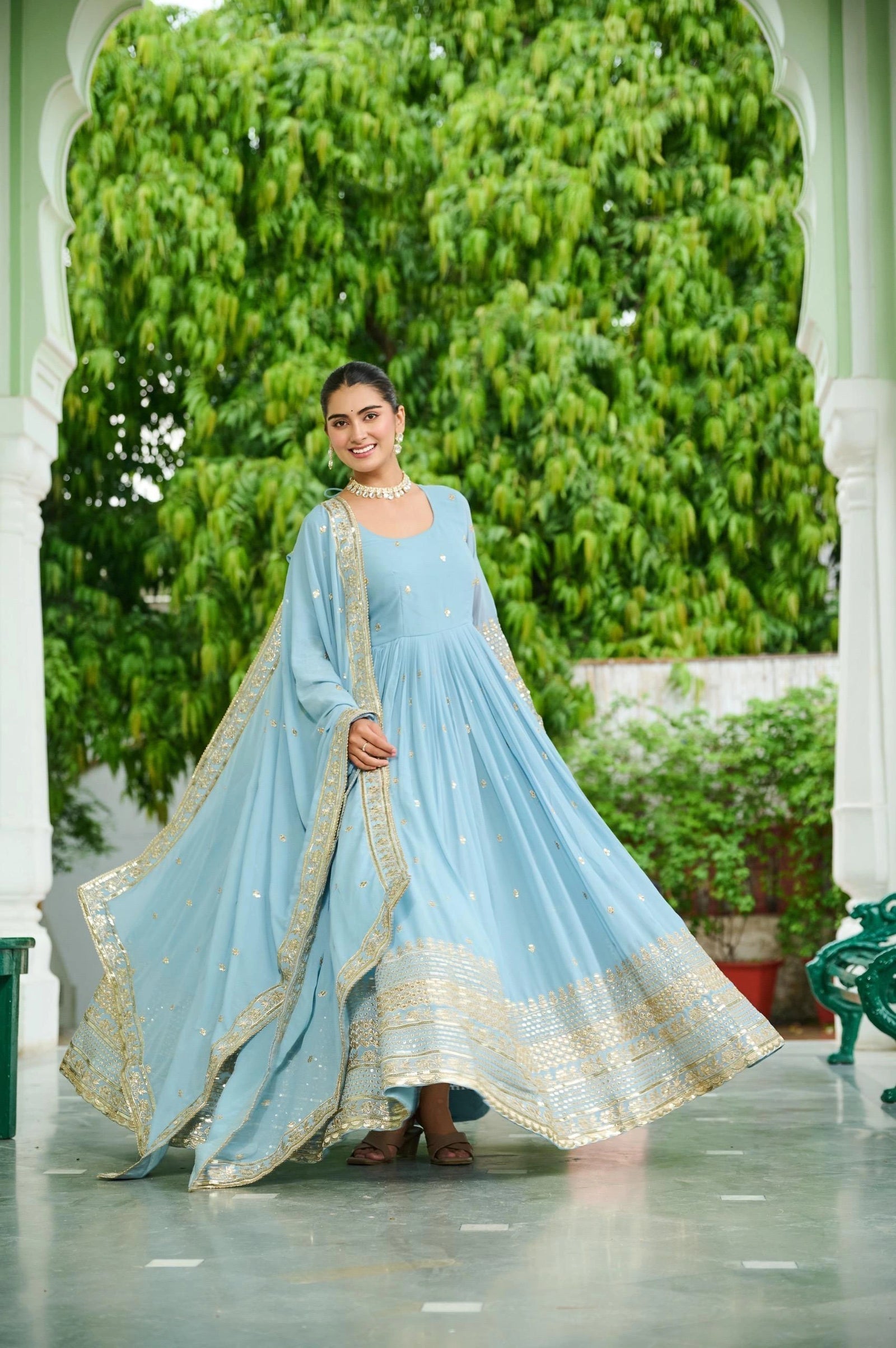 Woman in a light blue traditional outfit with gold details standing in front of greenery.