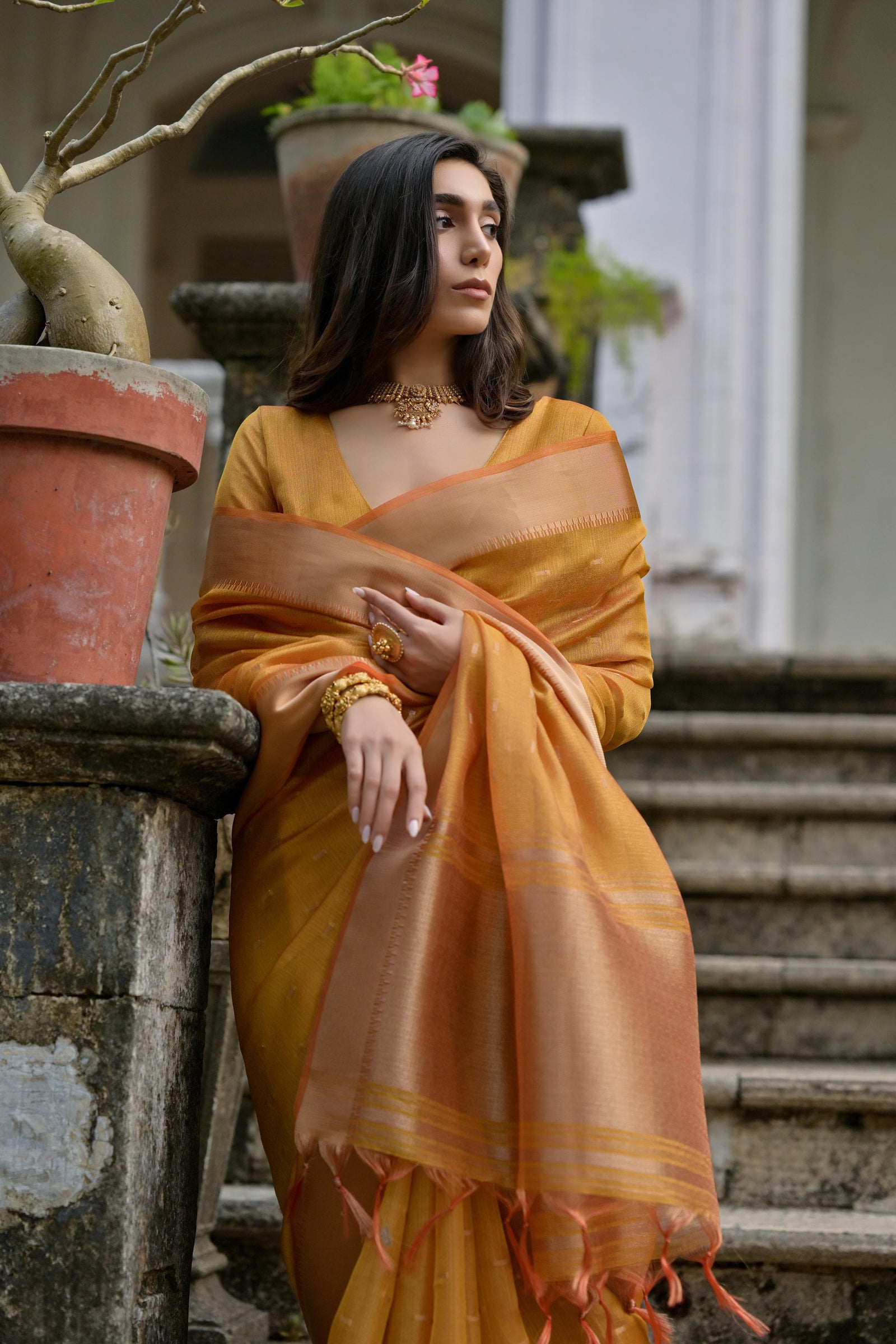 A woman models a stunning Golden Yellow Soft Silk Saree. The saree features subtle metallic motifs on the body and a wide, contrasting Copper/Rust Zari woven border. The lightweight fabric flows elegantly as she poses on a weathered stone staircase, wearing a delicate gold choker necklace.