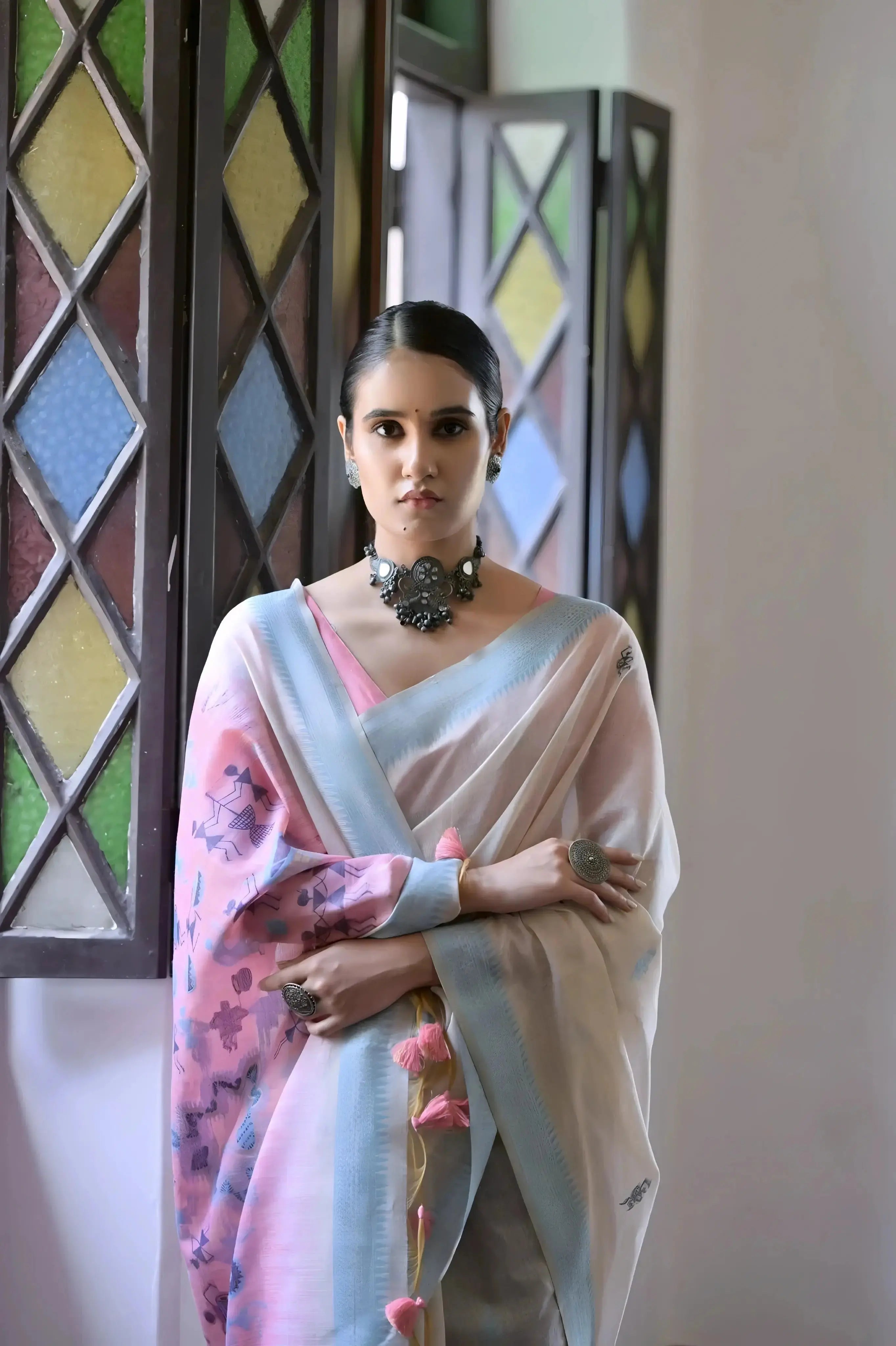 A woman models a graceful Off White Soft Muga Cotton Silk Saree. The saree has a light blue border, and the pallu features a wide pink panel decorated with traditional Warli-style figurative motifs in blue and purple. She wears a pink blouse and a dark oxidized choker necklace, posing indoors.