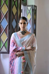 A woman models a graceful Off White Soft Muga Cotton Silk Saree. The saree has a light blue border, and the pallu features a wide pink panel decorated with traditional Warli-style figurative motifs in blue and purple. She wears a pink blouse and a dark oxidized choker necklace, posing indoors.