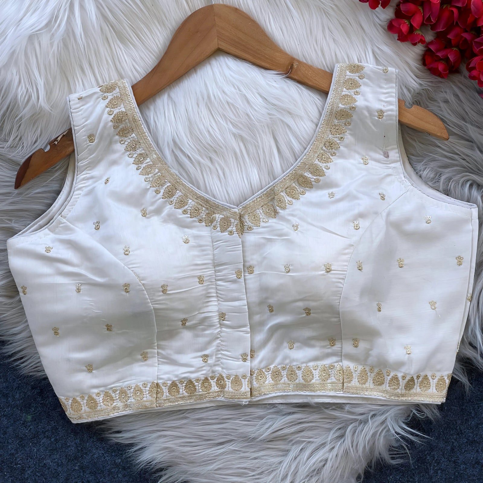 Ivory padded blouse with gold embroidery along the neckline and hem, featuring small gold embroidered motifs, displayed on a wooden hanger over a white fur surface with red artificial flowers in the background.