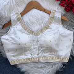 Ivory padded blouse with gold embroidery along the neckline and hem, featuring small gold embroidered motifs, displayed on a wooden hanger over a white fur surface with red artificial flowers in the background.