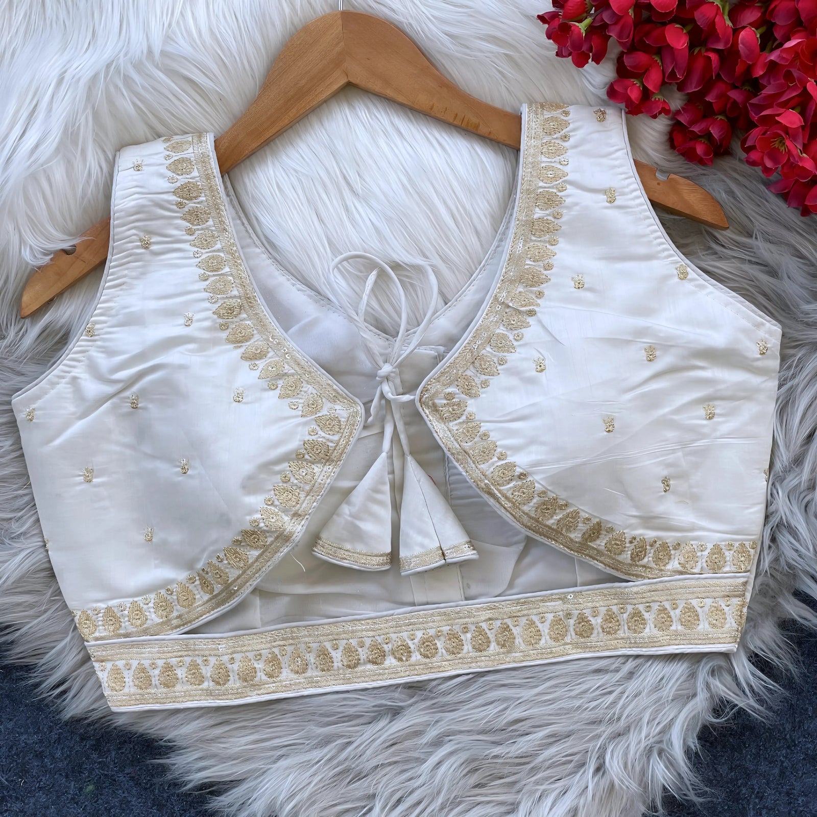 Ivory padded blouse with gold embroidery along the neckline and hem, featuring small gold embroidered motifs, displayed on a wooden hanger over a white fur surface with red artificial flowers in the background.