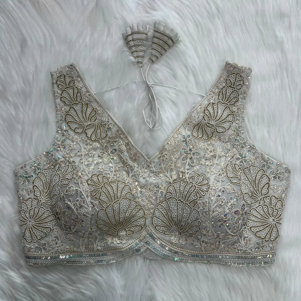 Ivory blouse featuring intricate gold sequin and thread embroidery with floral and shell motifs, shimmering bead embellishments, a deep V-neck, structured cups, and decorative tasseled tie-back detail, displayed flat on a white fur background.