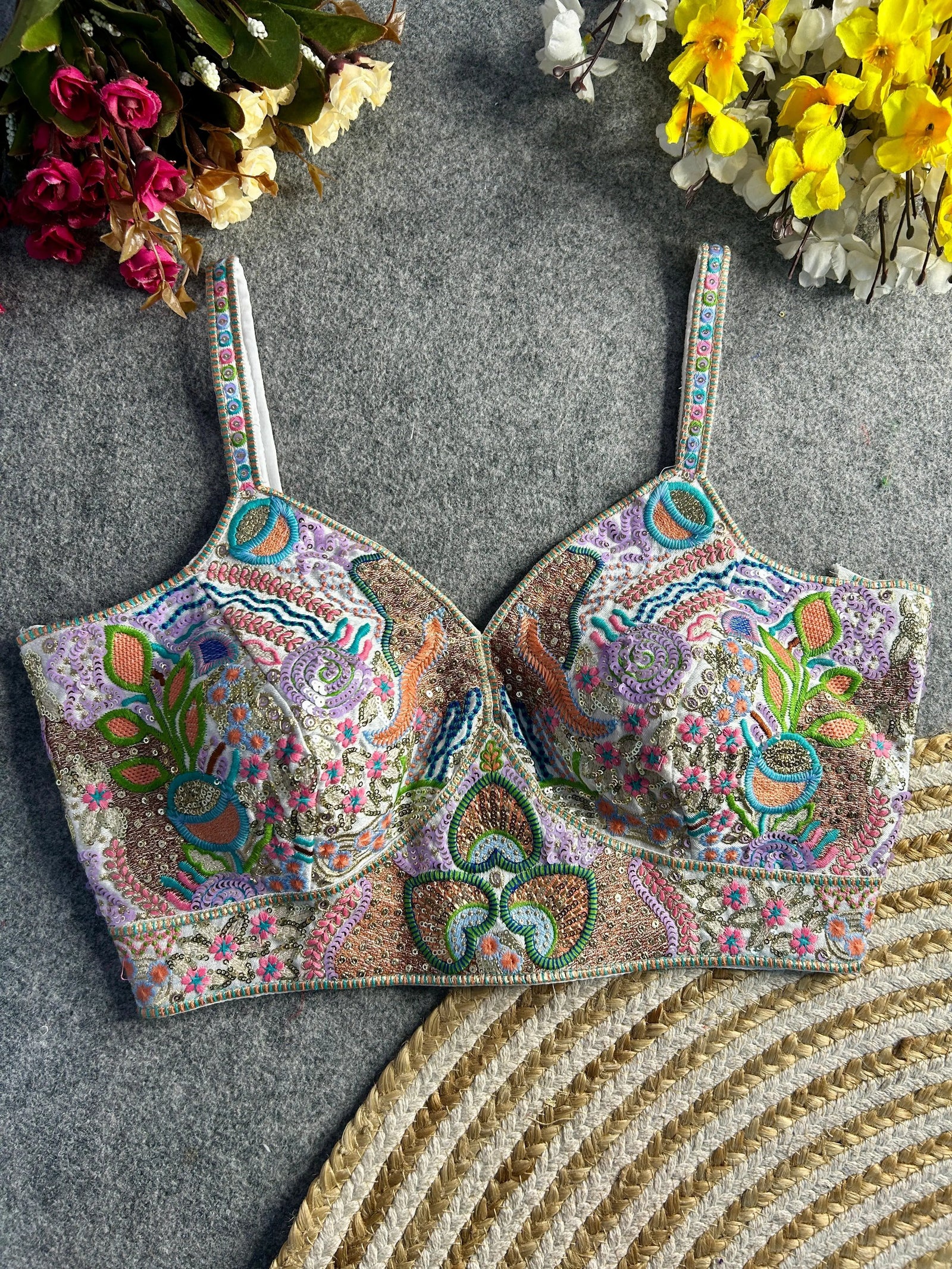 Traditional pastel bustier-style blouse featuring vibrant multicolor Madhubani-inspired embroidery with intricate floral, paisley, and geometric motifs, embellished with sequins and beads, structured cups, and adjustable straps, displayed flat on a textured grey surface with artificial pink, yellow, and white flowers in the corners.
