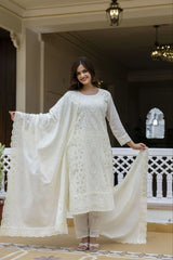 Woman in a white embroidered outfit holding a matching dupatta in an elegant indoor setting.