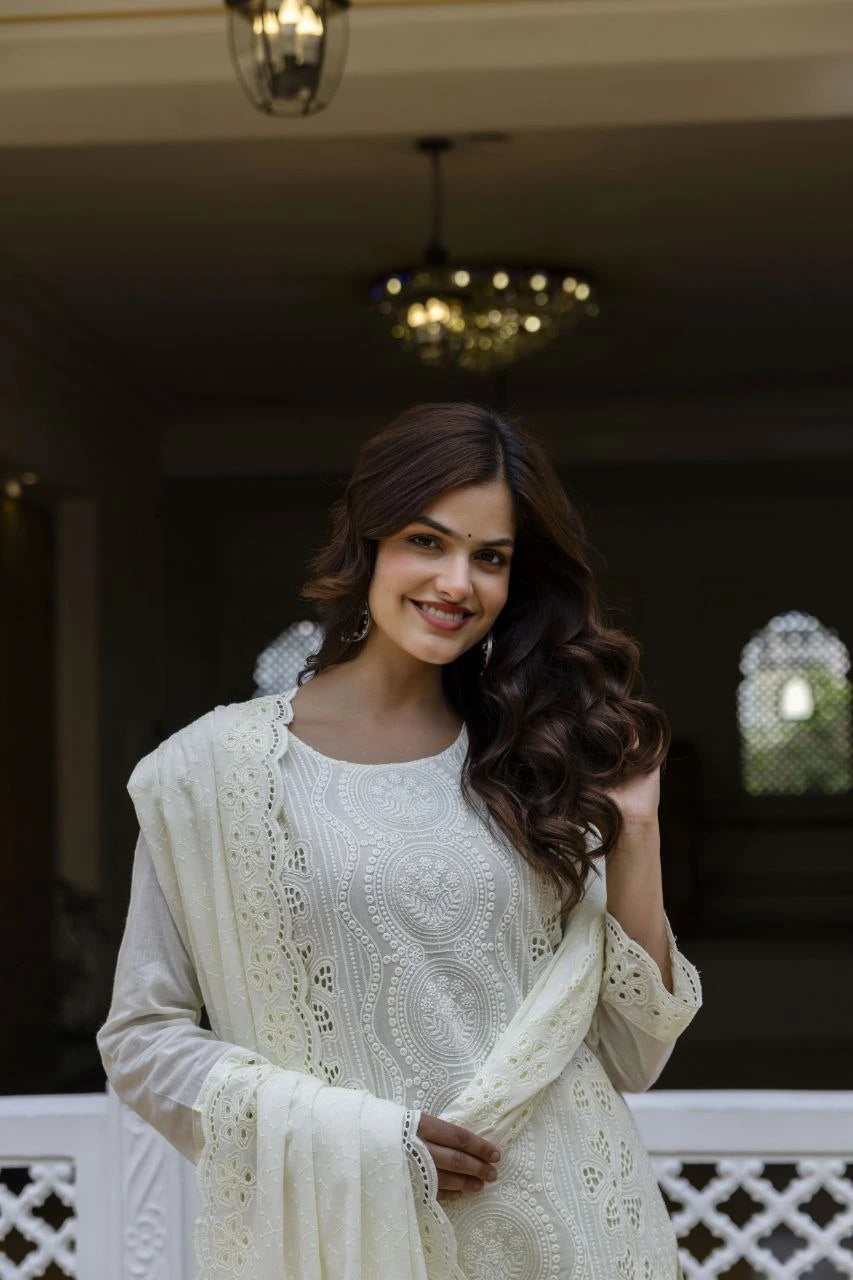 Woman in a white traditional outfit with intricate patterns, standing indoors.