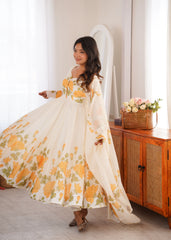Woman in a floral dress standing in a room with a wooden cabinet and flowers.