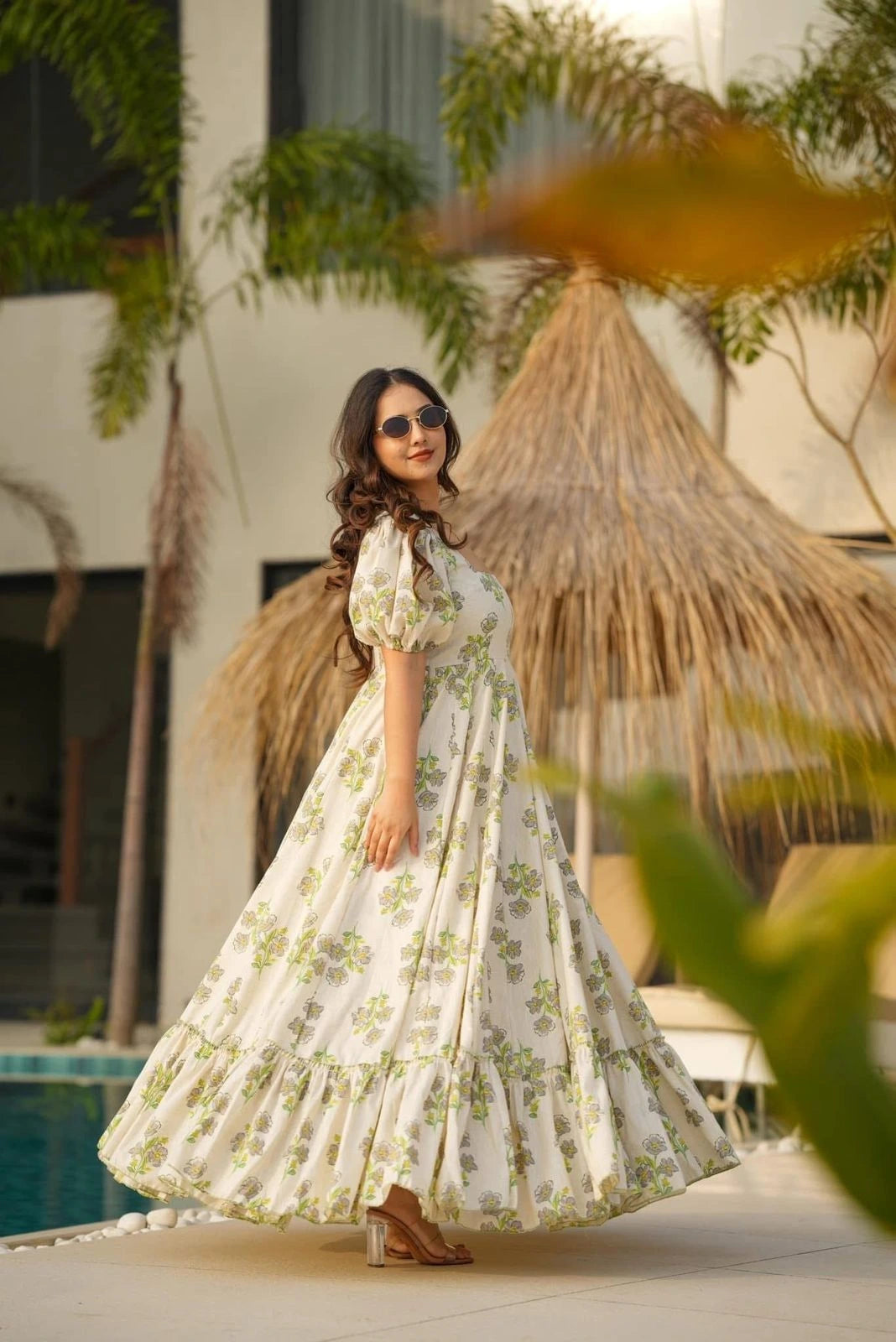 Woman in a floral dress standing by a poolside with plants and a thatched umbrella in the background.