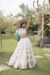 Elegant white lehenga choli featuring a sleeveless mirror work blouse and a flared skirt with bold golden polka dot motifs.