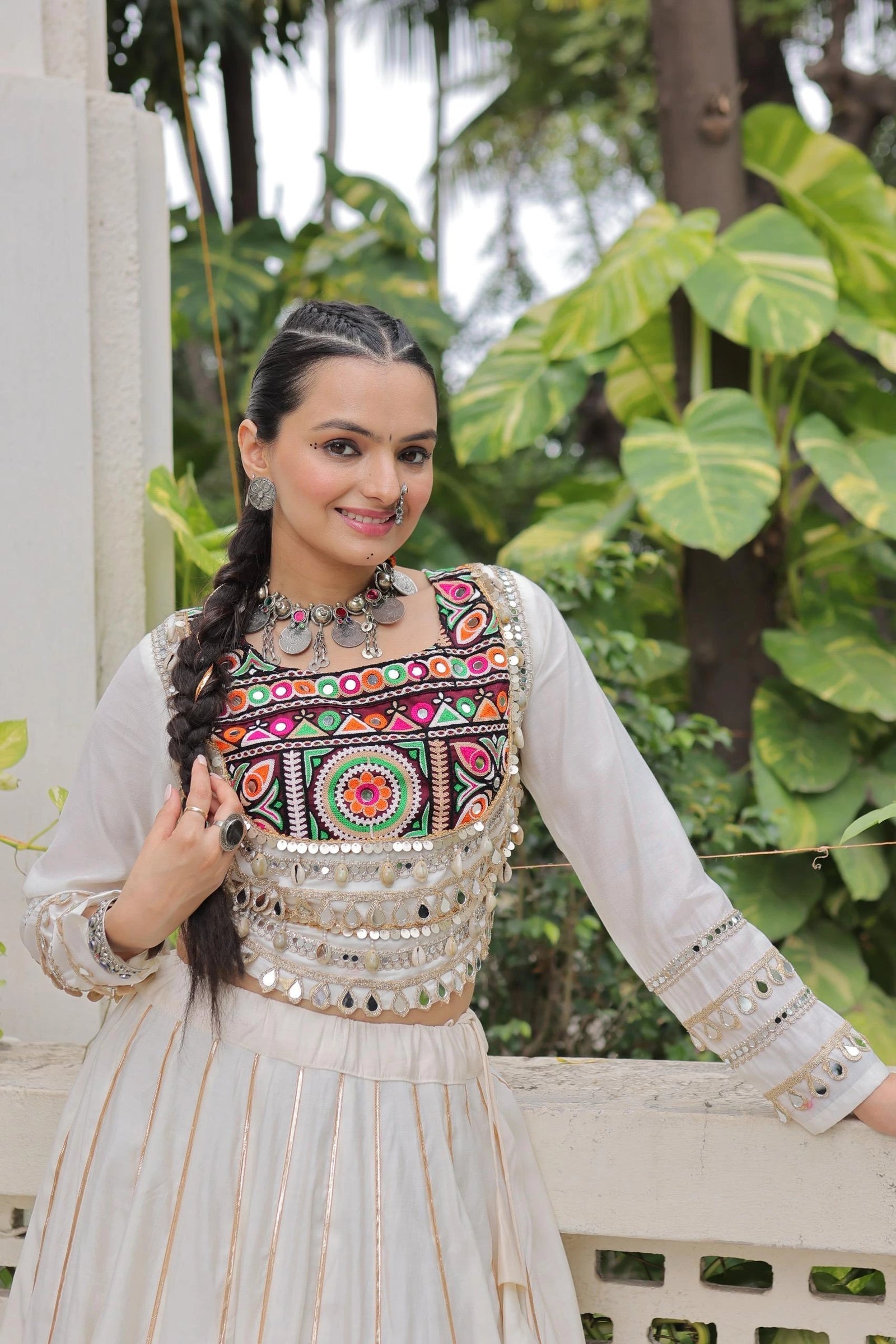Woman wearing a traditional embroidered top with a white outfit, standing outdoors with greenery in the background.
