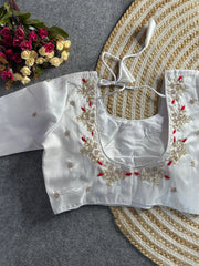 A pristine white raw silk blouse featuring a wide, rounded back neckline. The edges are decorated with elegant hand-embroidery of gold and pearl floral sprigs, accented by vibrant red threadwork. It includes matching white tie-back cords with delicate tassels, and small pearl floral motifs are scattered across the back and sleeves.