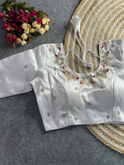The front of the white silk blouse showcases a curved V-neckline with the same intricate pearl and gold floral embroidery and red accents. The garment is tailored with structured princess-line seams and is adorned with scattered embroidered pearl buttis. The blouse is laid flat on a grey textured background next to a round jute mat and a bouquet of roses.