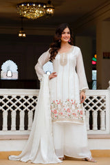 Woman in a white traditional outfit with floral design standing in an elegant indoor setting.