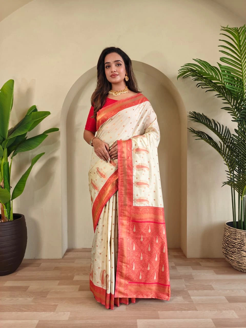 Woman in a white and red saree standing in a room with plants.
