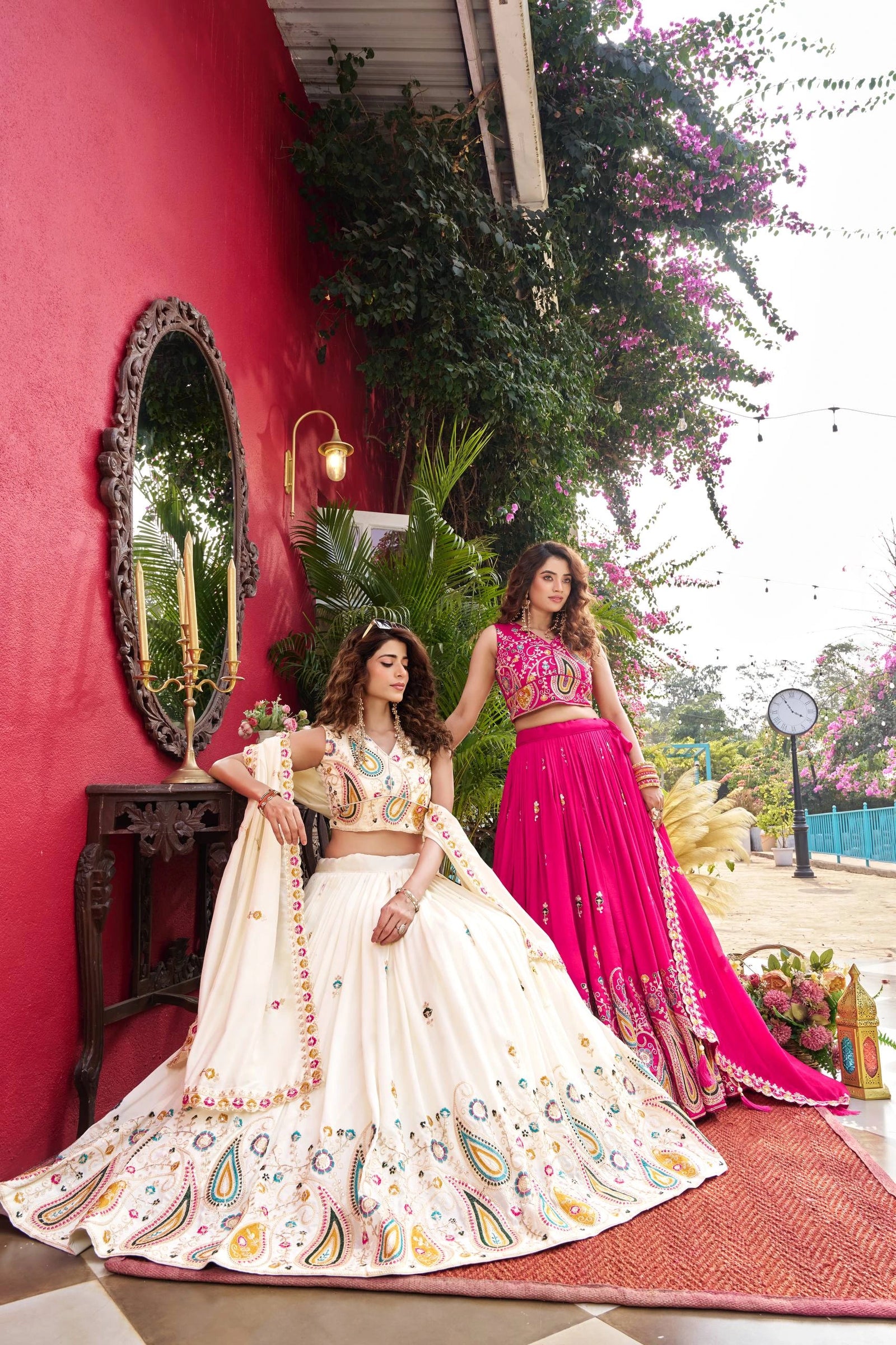 Two women in traditional outfits standing in front of a red wall with greenery.