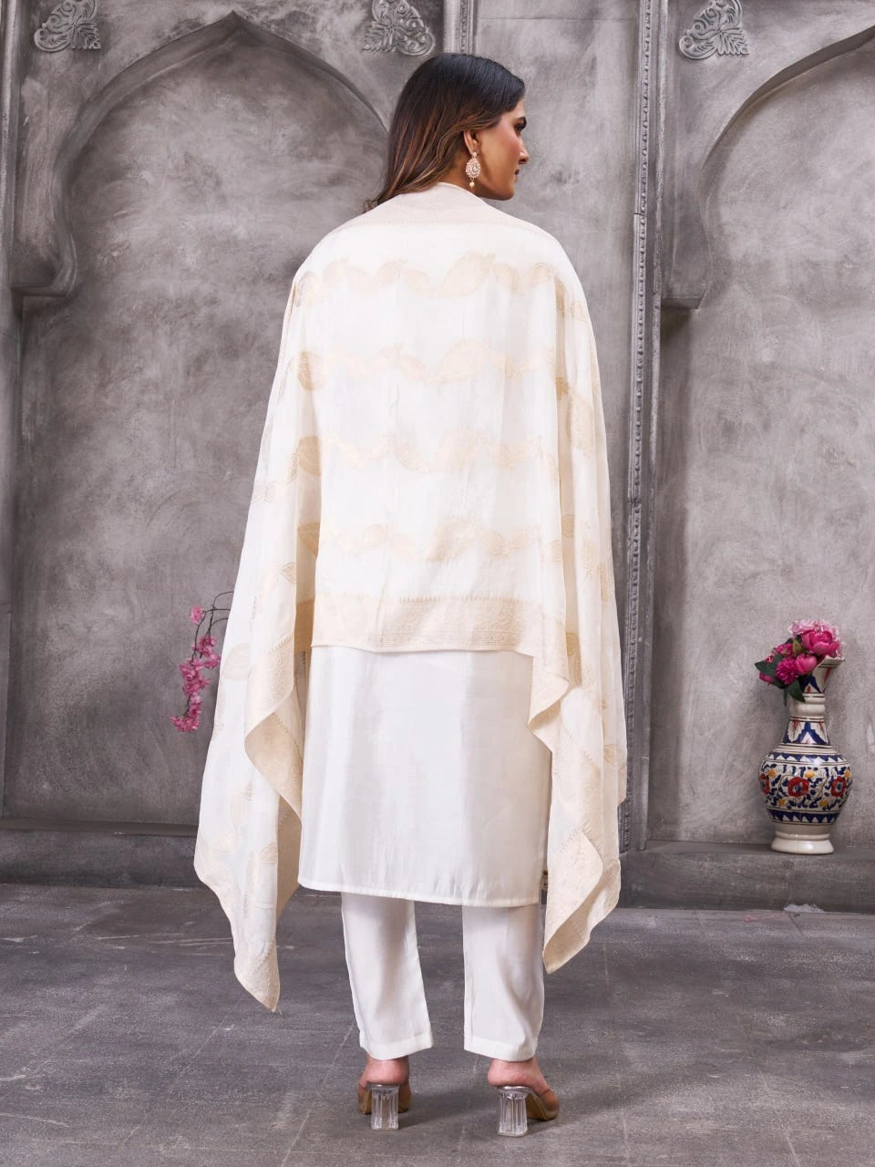 Woman wearing a cream-colored outfit with a long drape against a gray wall.