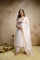 Woman in traditional white outfit with floral embroidery standing against a textured beige wall.