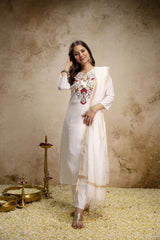 Woman in a white traditional outfit with intricate patterns, standing against a textured beige wall.