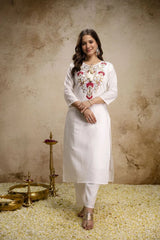 Woman wearing a white embroidered kurta with gold jewelry against a textured beige background