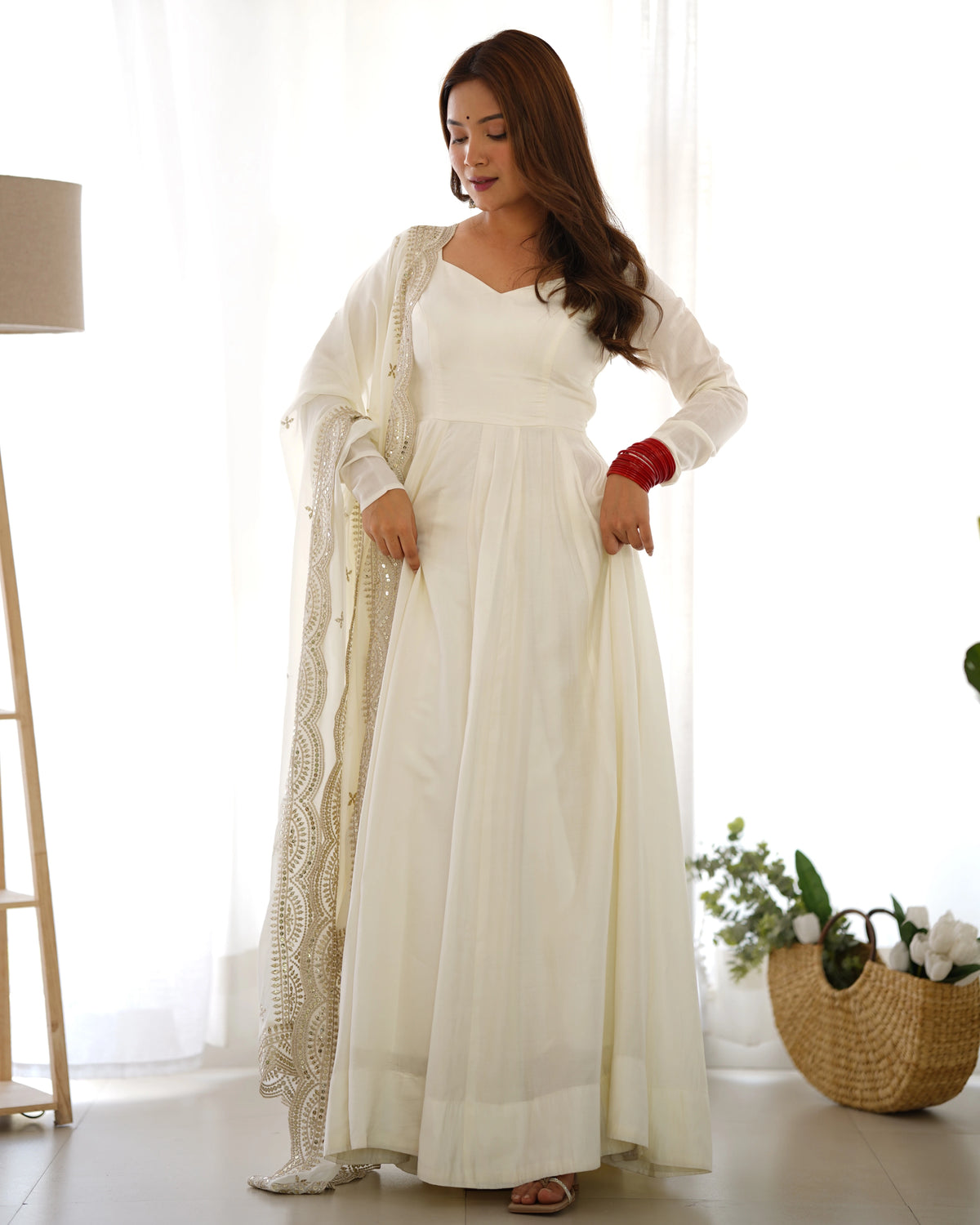 Woman in a white dress with a shawl standing in a bright room with a lamp and plant.