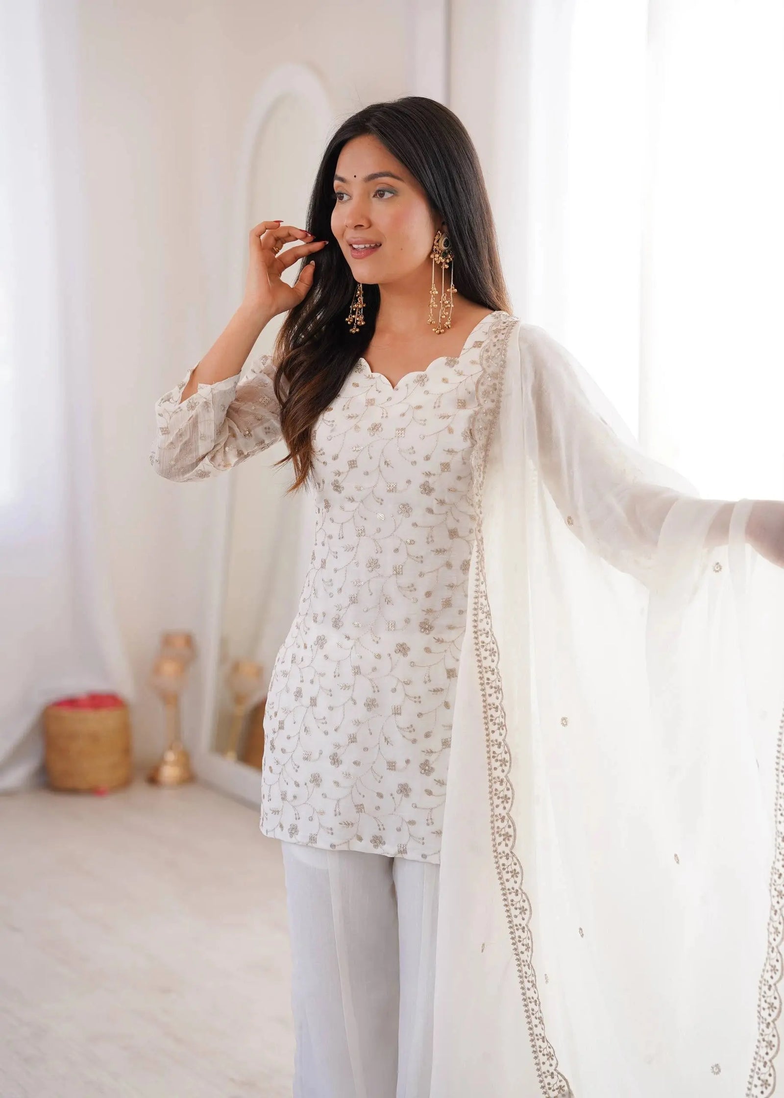 Woman wearing a white embroidered outfit with a sheer dupatta in a softly lit room.