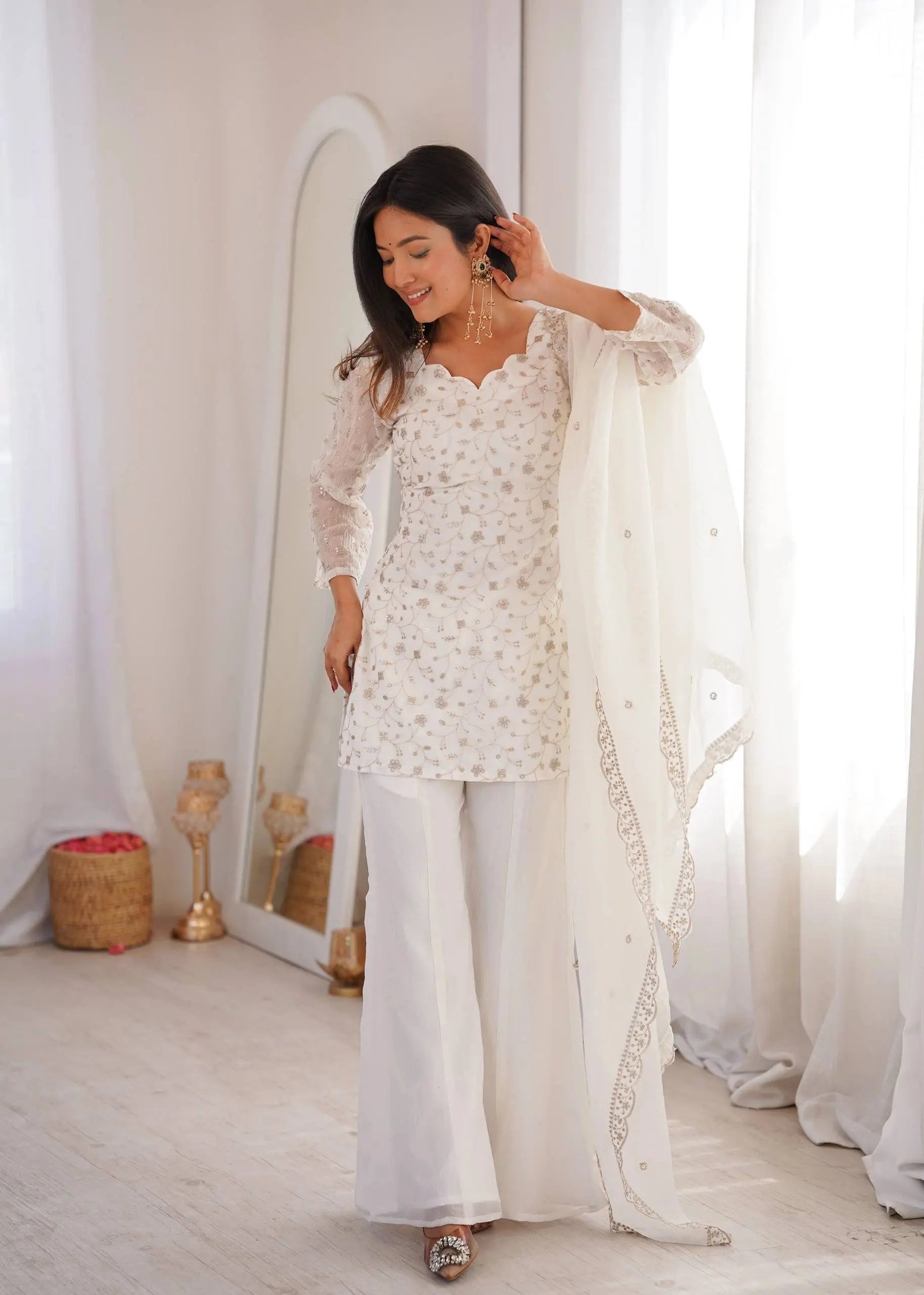 Woman in a white traditional outfit standing in a bright room with white curtains.