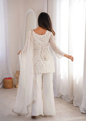 Woman in a white traditional outfit standing in a bright room with sheer curtains.
