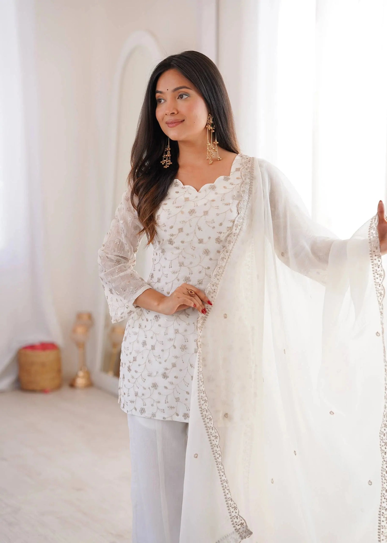 Woman wearing a white embroidered outfit with a sheer dupatta in a softly lit room.