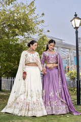 Two women in traditional embroidered lehengas standing outdoors with greenery and a lamp post in the background.