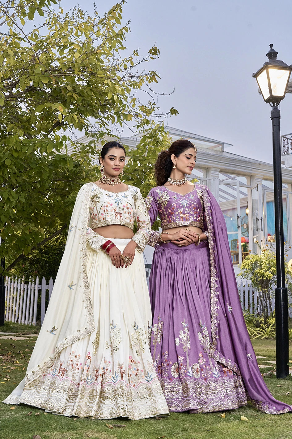 Two women in traditional outfits standing outdoors with greenery and a building in the background.