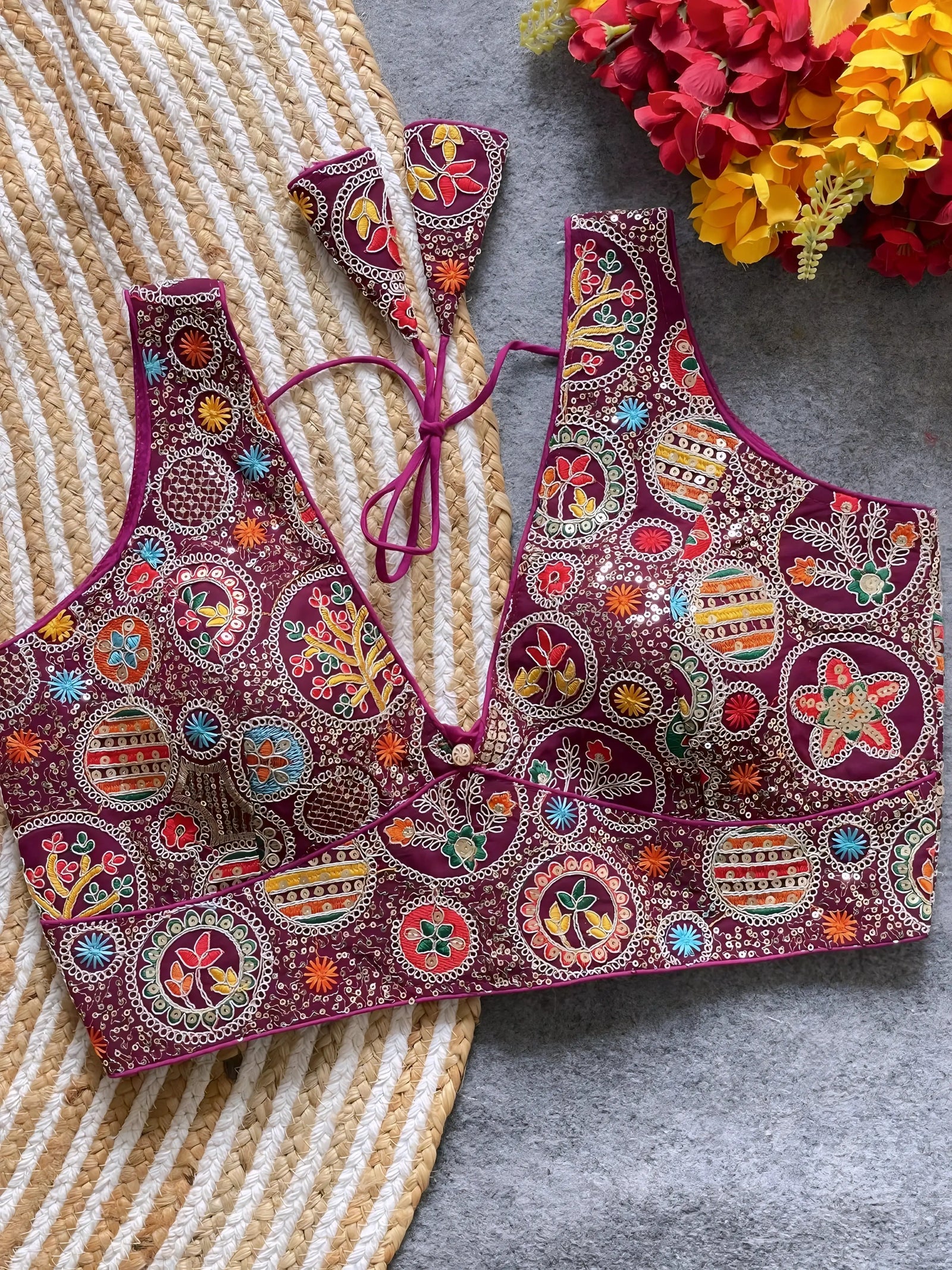 Traditional maroon blouse featuring vibrant multicolor thread embroidery and mirror work in circular floral and geometric patterns, with a deep V-neck and tie-back detail, displayed flat on a textured mat with artificial red and yellow flowers in the corner.