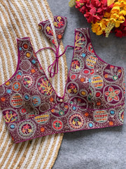 Traditional maroon blouse featuring vibrant multicolor thread embroidery and mirror work in circular floral and geometric patterns, with a deep V-neck and tie-back detail, displayed flat on a textured mat with artificial red and yellow flowers in the corner.