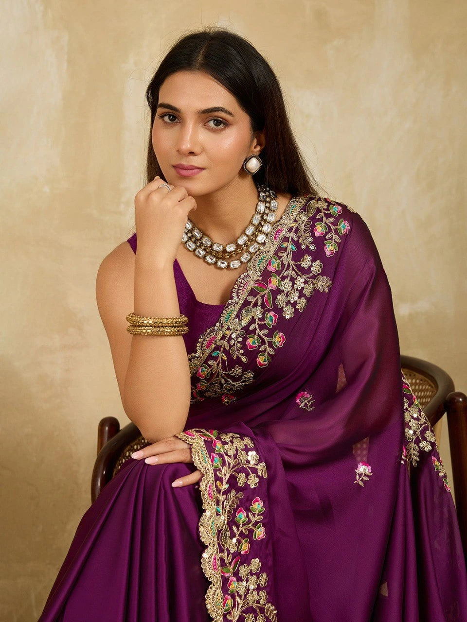 Woman wearing a purple saree with intricate gold embroidery against a beige background